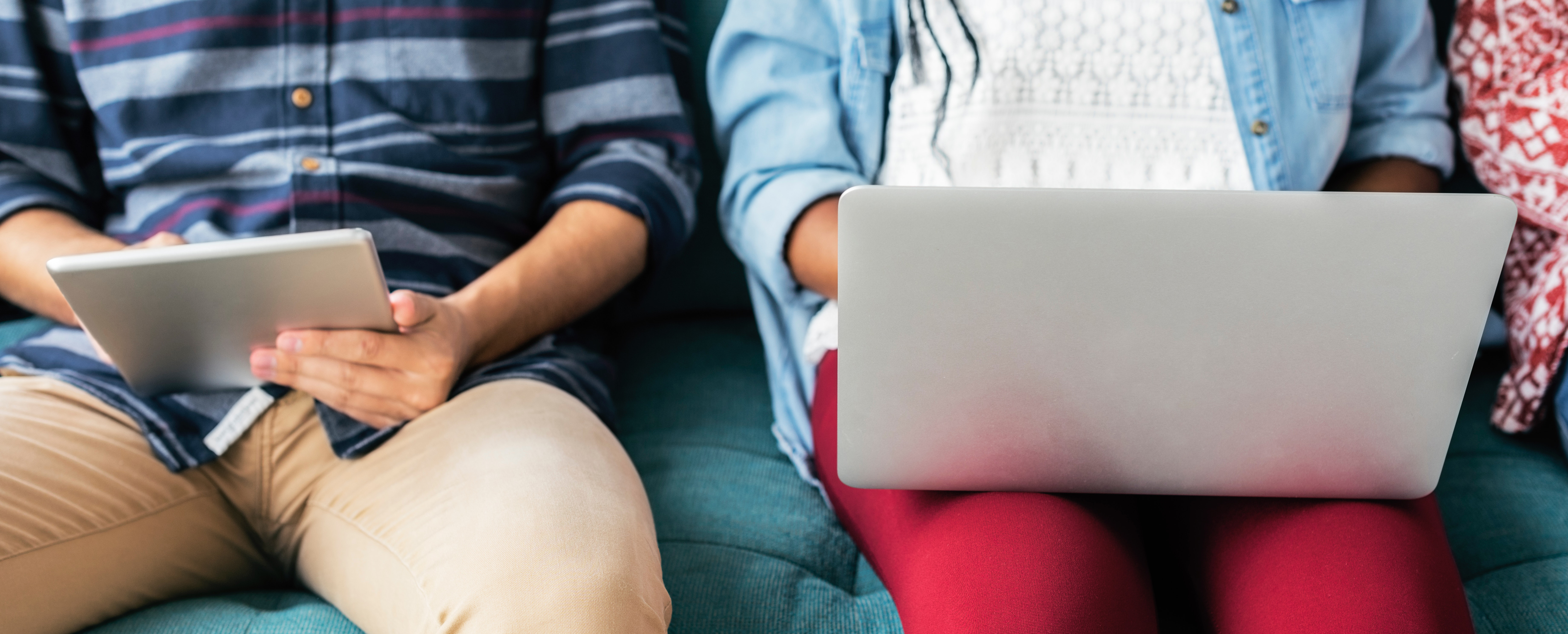two students sitting on a couch; one works on a laptop while the other works on a tablet