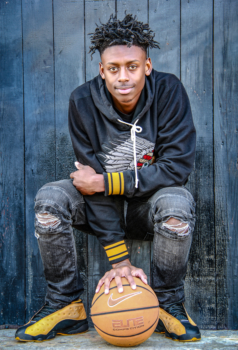 Photo of young man in ripped jeans sitting holding a basketball in place on the ground. Man is looking into camera.